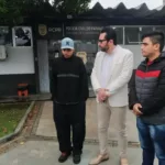 Imagem colorida mostra frentista vítima de racismo, vestido de moletom preto e boné azul, ao lado de seu advogado, de terno claro, e outro homem, de casaco, em frente a delegacia em Curitiba - Metrópoles